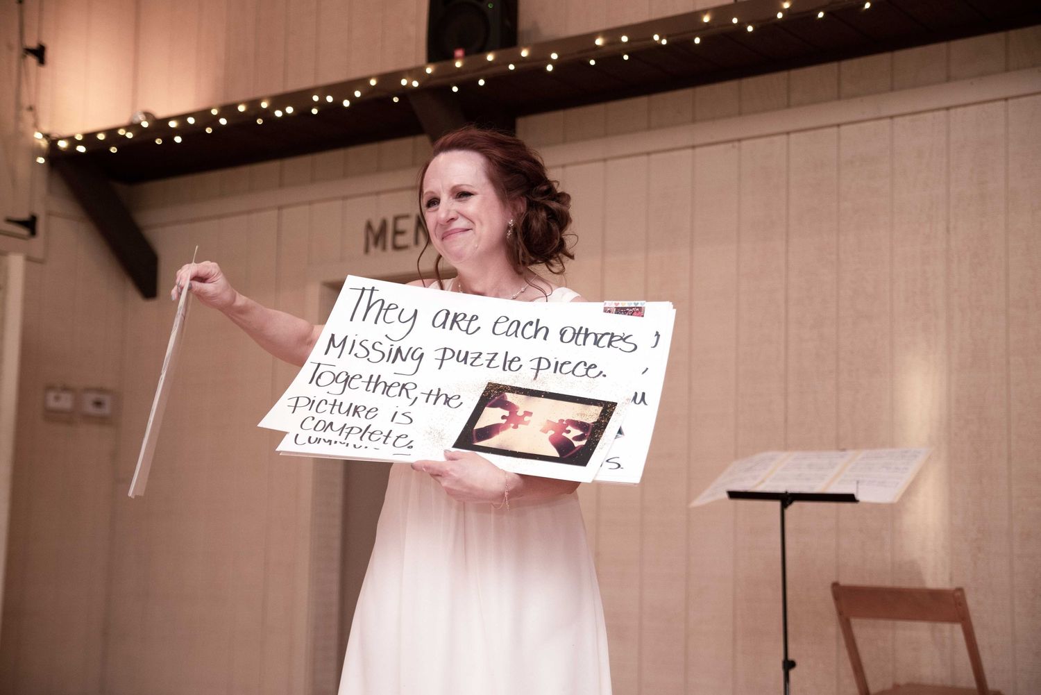 Woman in white dress holds a handwritten sign with a photo at a wedding reception venue.