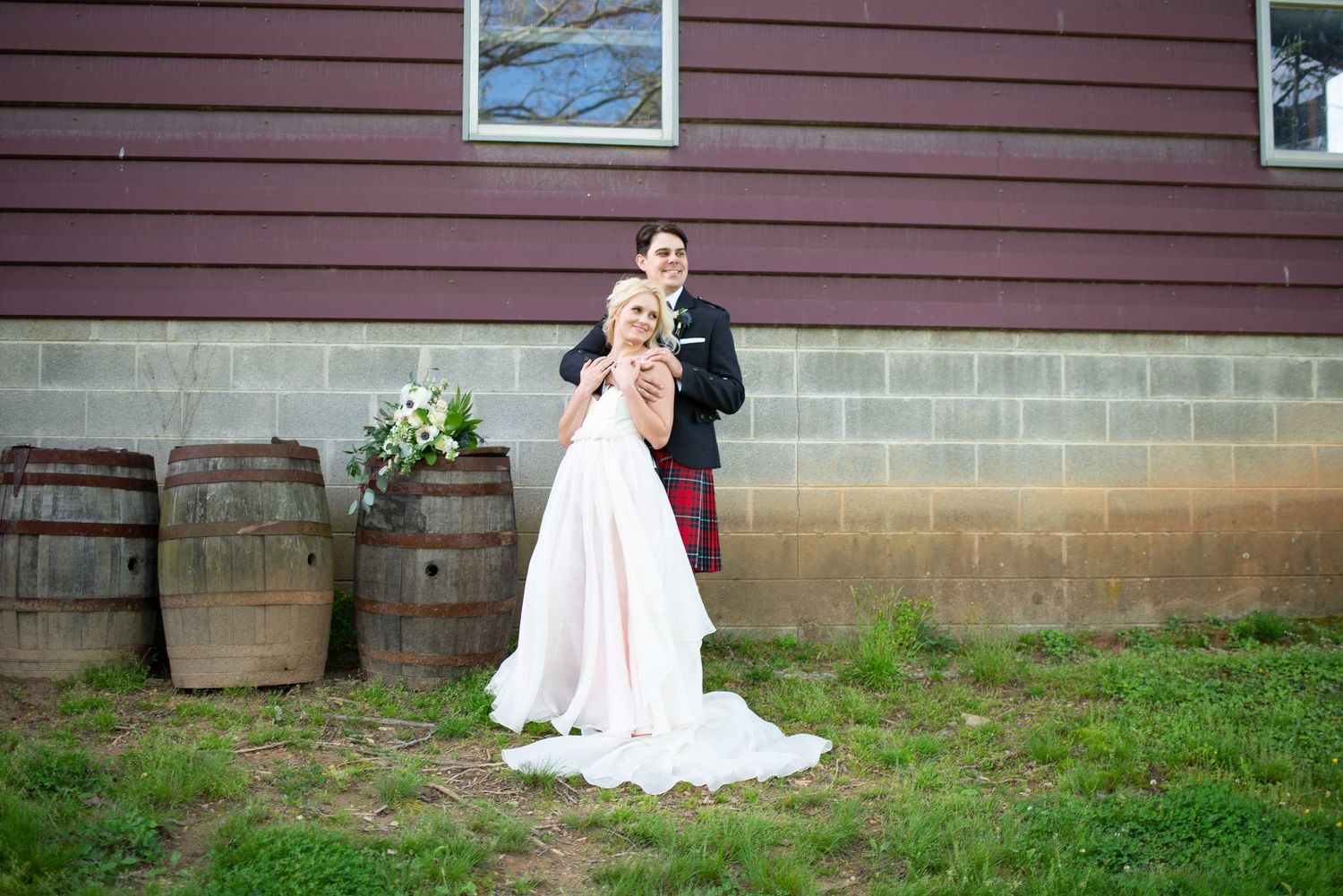 Bride and groom posing outdoors beside rustic wooden barrels and a red barn wall on their wedding day.