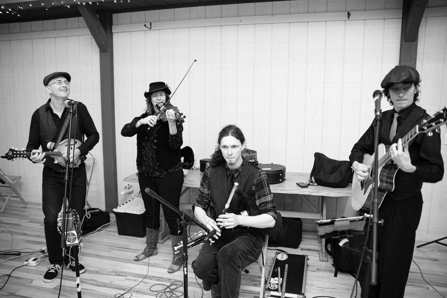Black and white photo of a four-piece folk band playing violin, flute, and guitars at an outdoor wooden venue.