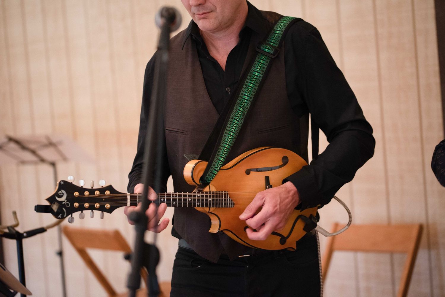 Musician playing a mandolin with a green strap, wearing all black, performing at an indoor event.