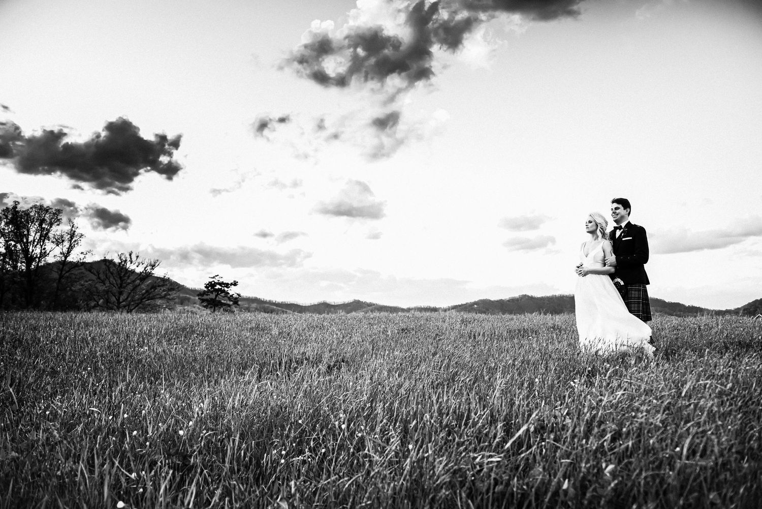 Black and white photo of a bride and groom embracing in a vast grassy field with rolling hills and dramatic cloudy sky.