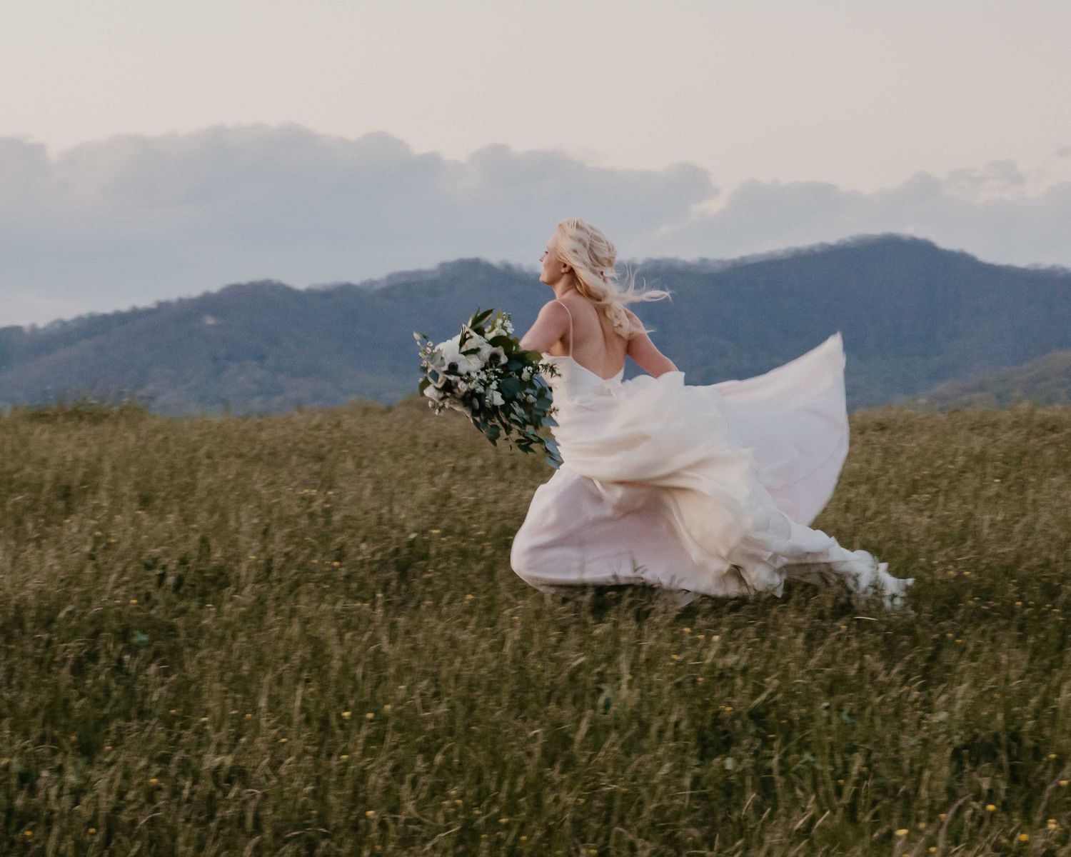 Bride in flowing white gown running through mountain meadow, hair blowing in wind, holding wildflower bouquet.