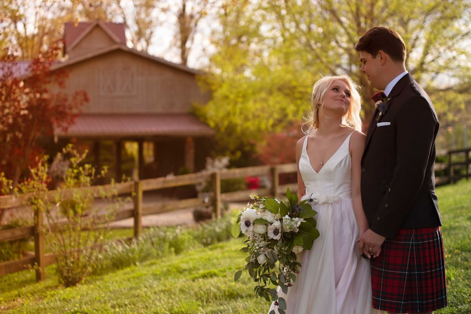 Bride and groom holding hands in a garden, she holds a white bouquet, he wears a red tartan kilt, rustic barn backdrop.