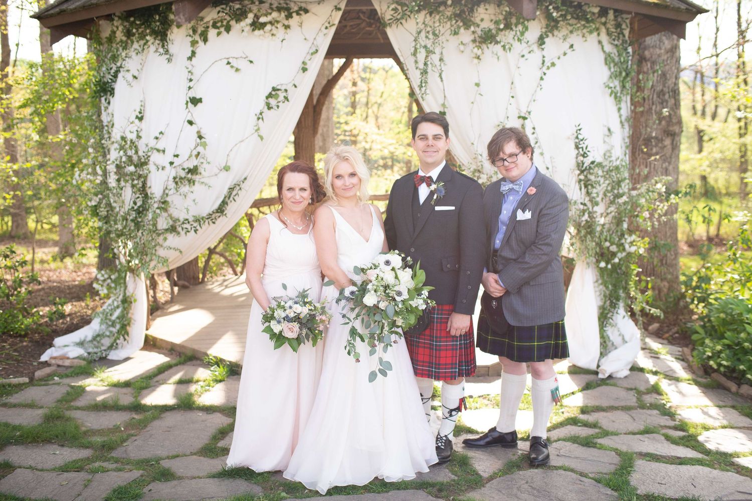 Wedding party poses under floral gazebo, bride in white gown, bridesmaid in pink, groom in tux, groomsman in kilt.