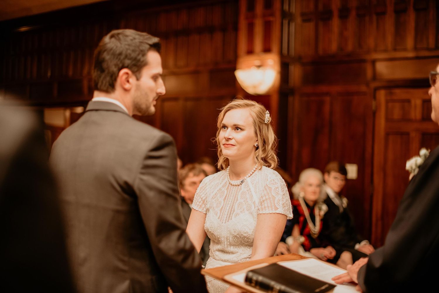 Bride smiling at groom during wedding ceremony in elegant wood-paneled venue with guests seated in background.