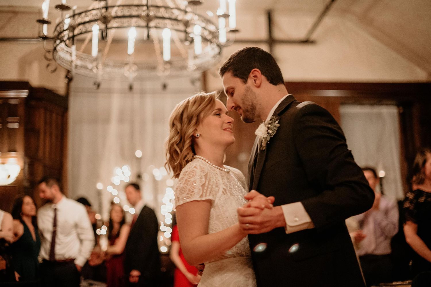 Bride and groom share first dance under chandelier with sparklers glowing in background at elegant wedding reception.