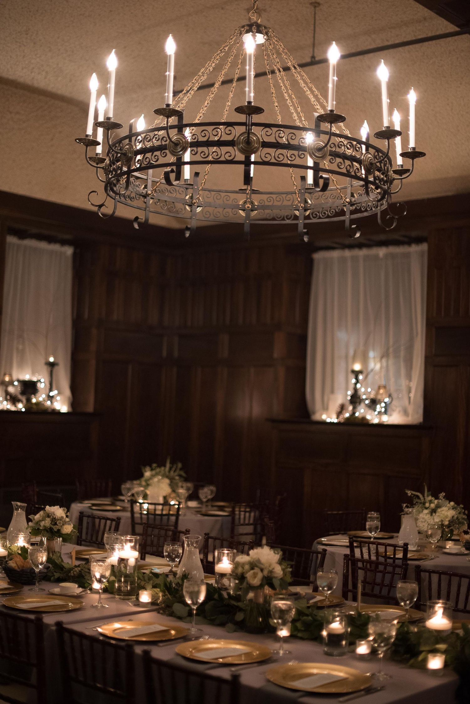 Elegant wedding reception with wrought iron chandelier, candlelit tables, gold chargers, and floral centerpieces in a dark wood room.