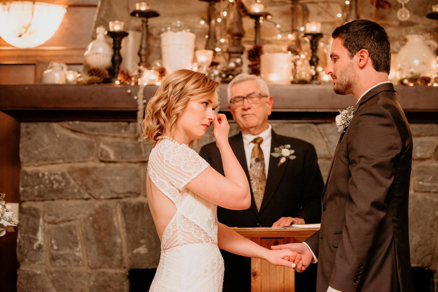 Bride wiping tears during emotional indoor wedding ceremony with groom and officiant at stone fireplace.