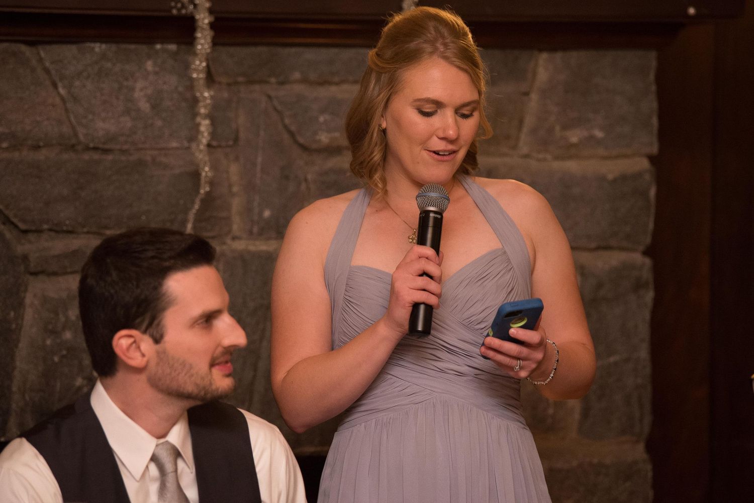 A bridesmaid in a gray dress gives a speech holding a microphone and phone at a wedding reception.