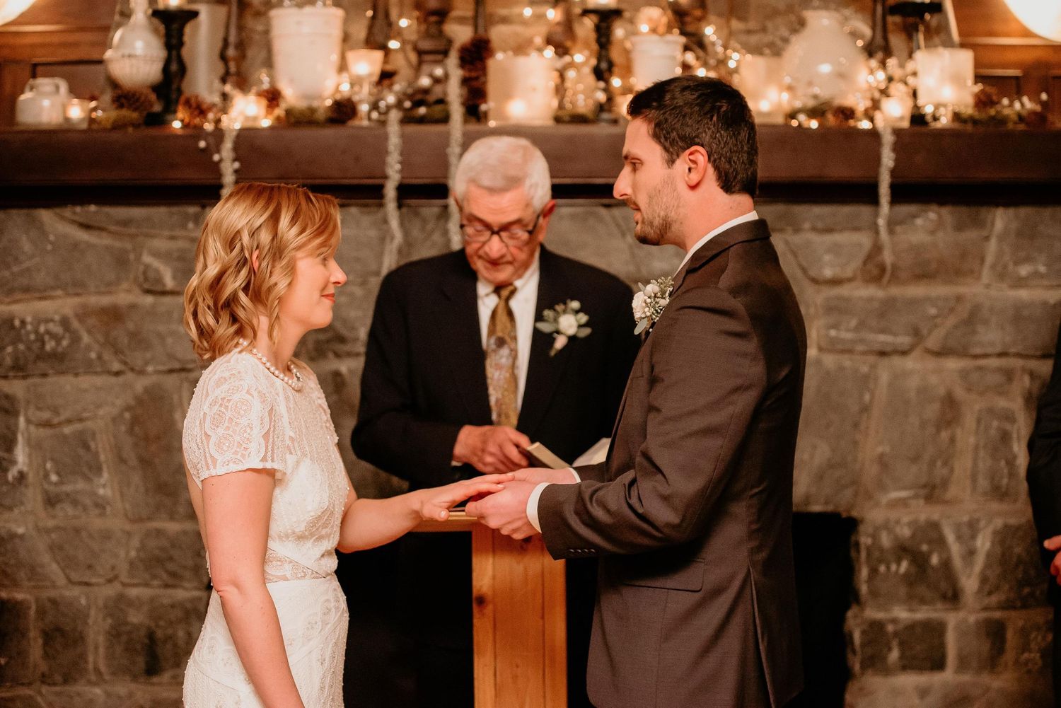Couple exchanging rings during intimate wedding ceremony before stone fireplace with officiant.