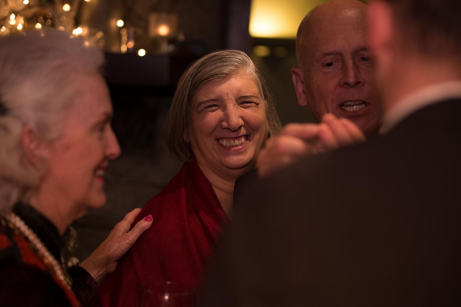 Three people socializing at an indoor evening event, woman in red top smiling warmly in conversation.