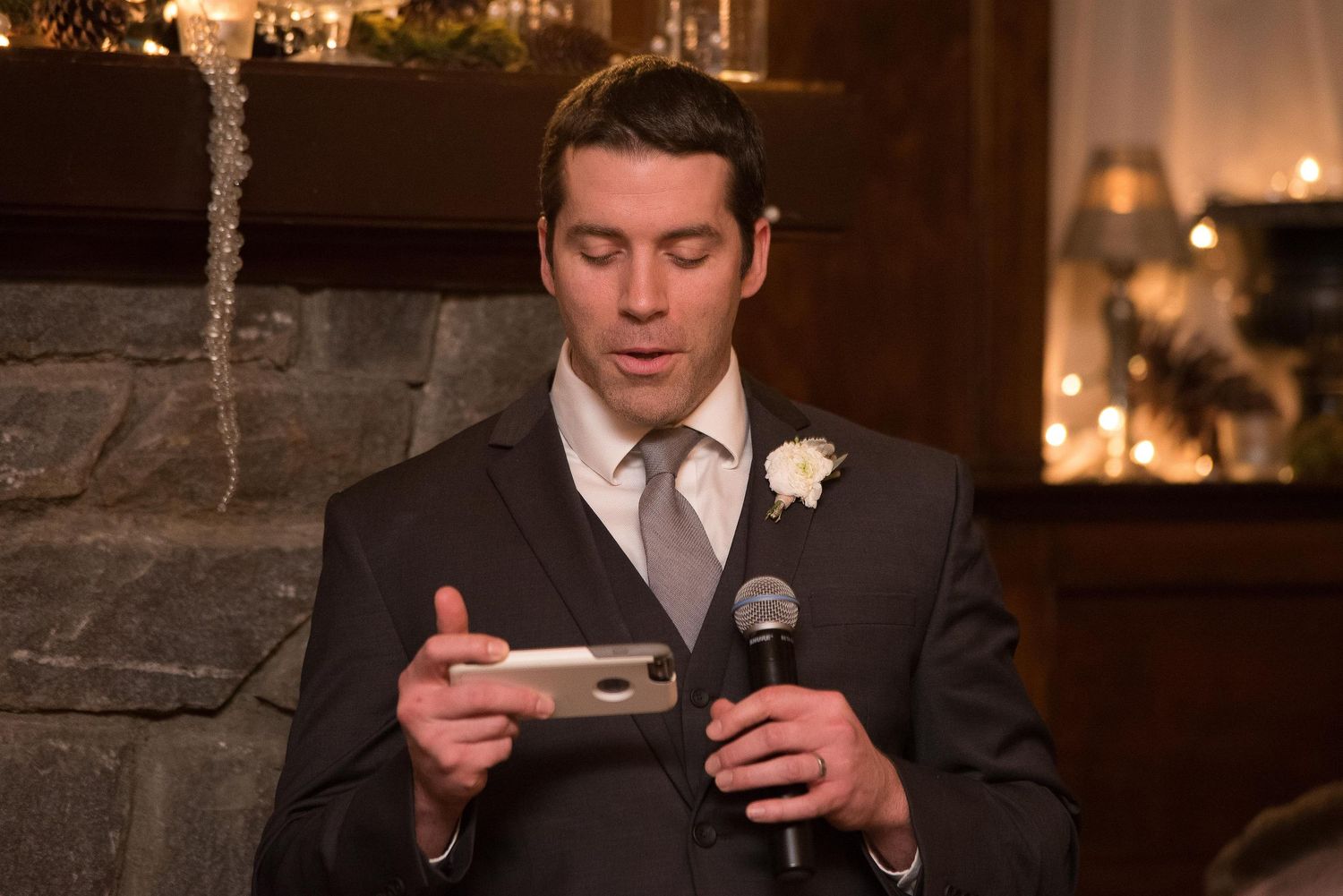 Man in suit with boutonniere reads from phone while speaking into microphone at wedding reception.