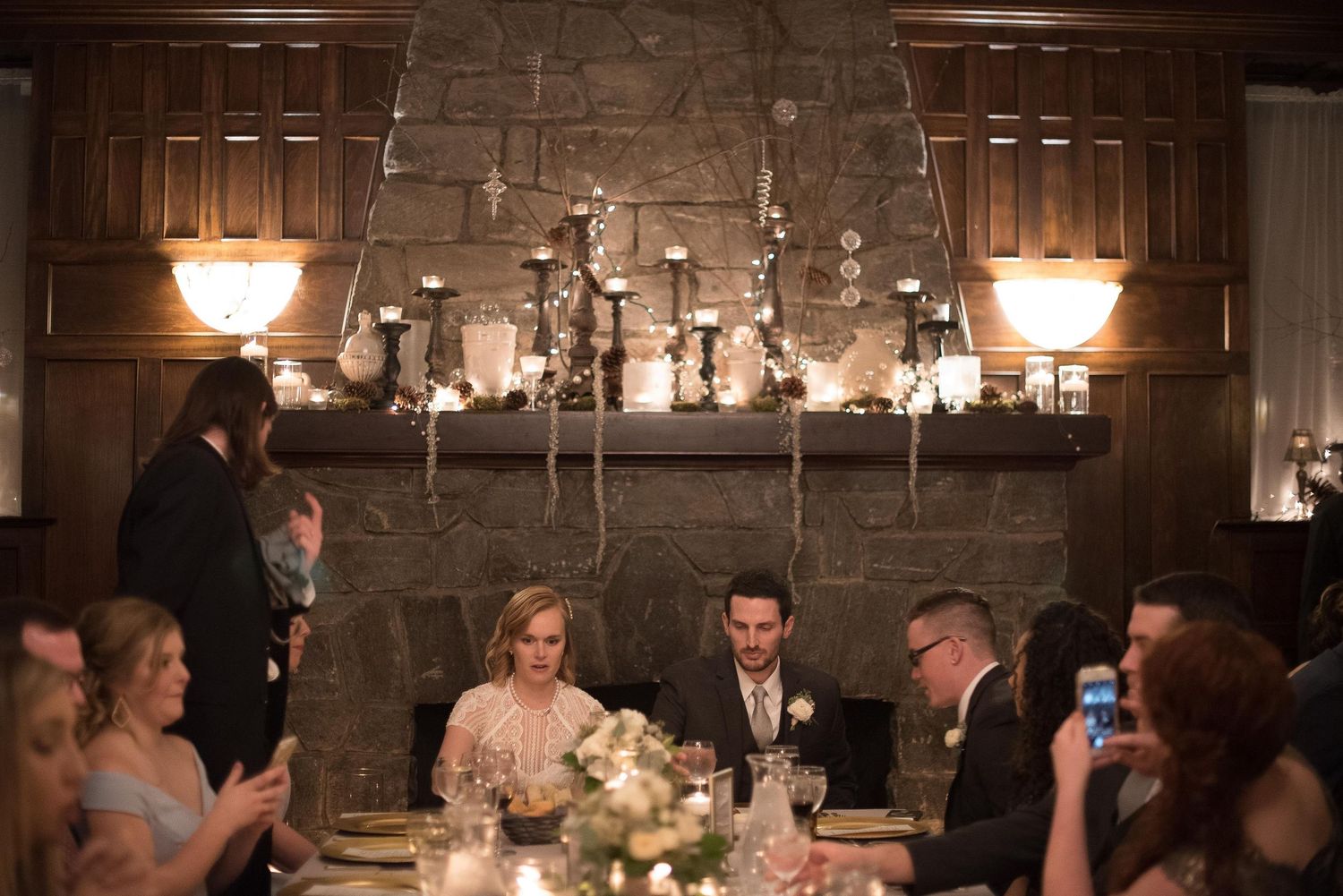 Bride and groom seated at head table during wedding reception with stone fireplace backdrop and candlelit ambiance.