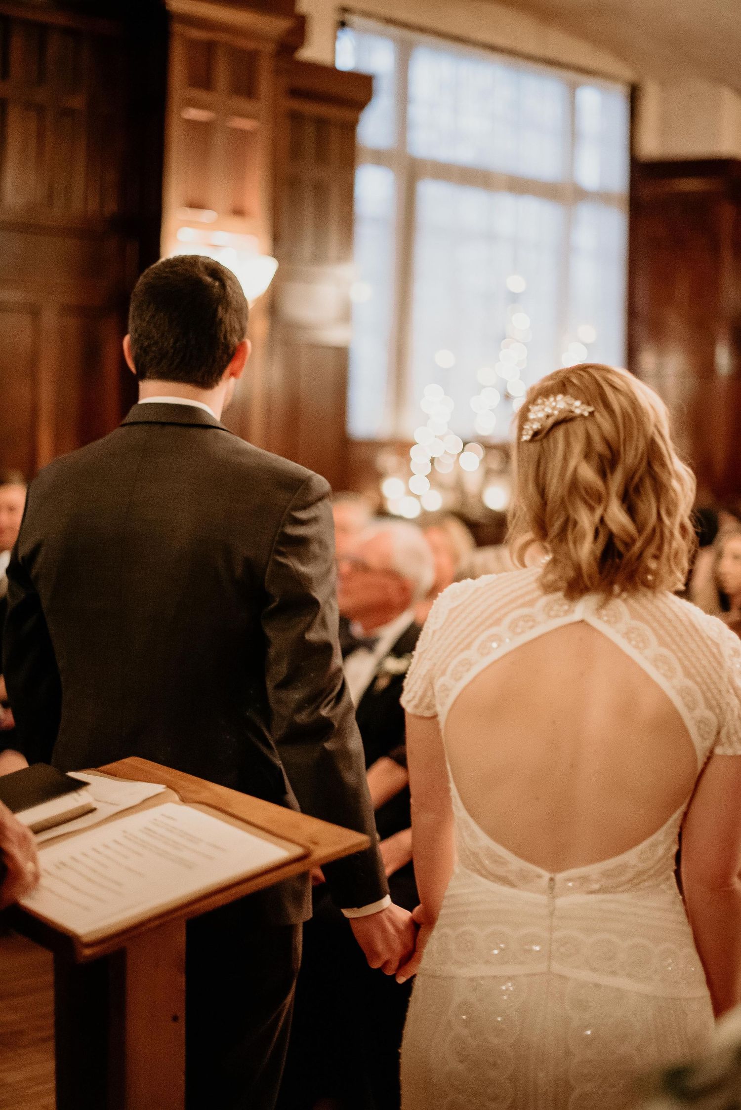 Bride and groom holding hands at wedding altar, bride in open-back lace gown with hair accessory.