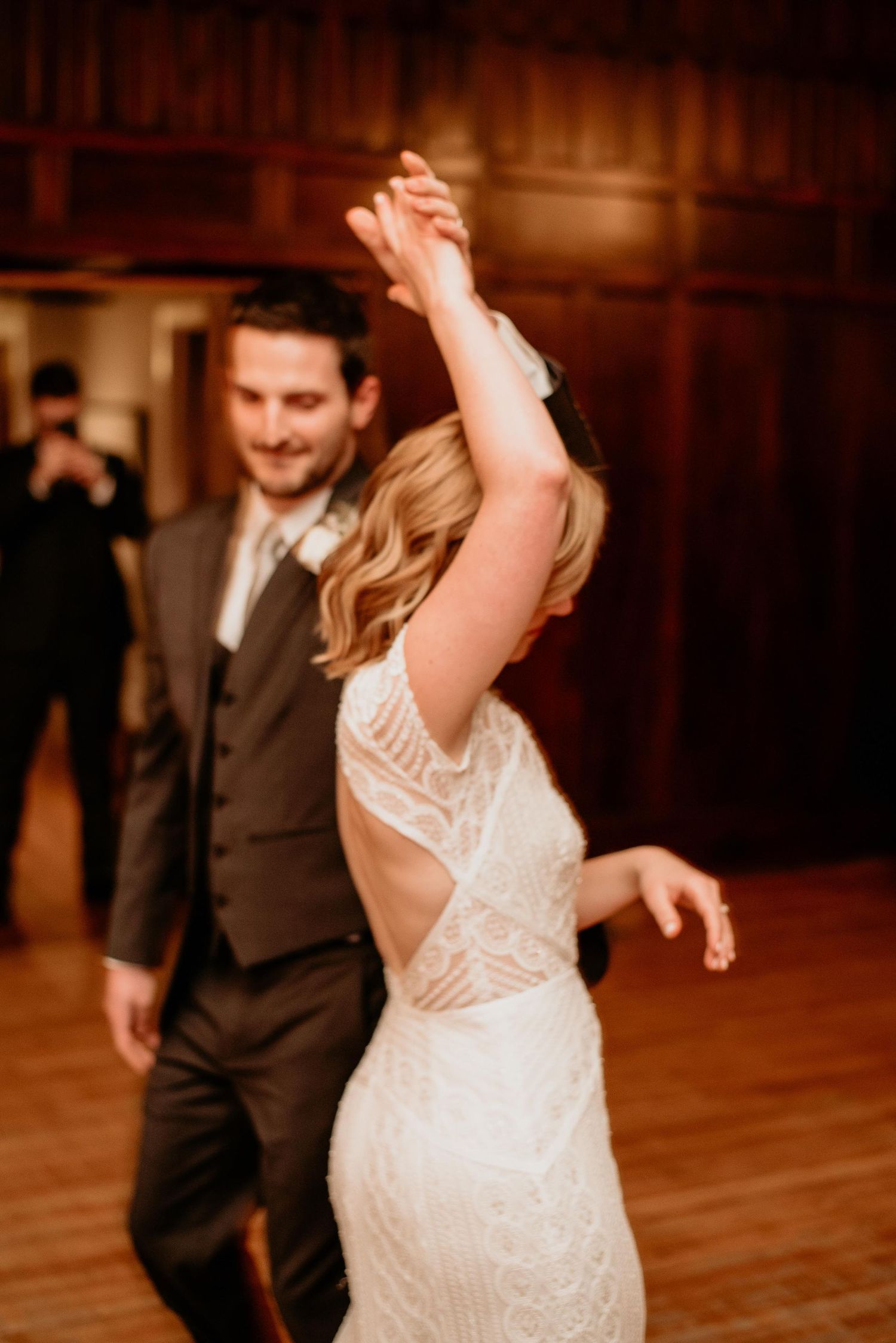Bride in lace backless gown dancing with groom in dark suit during elegant wedding reception.