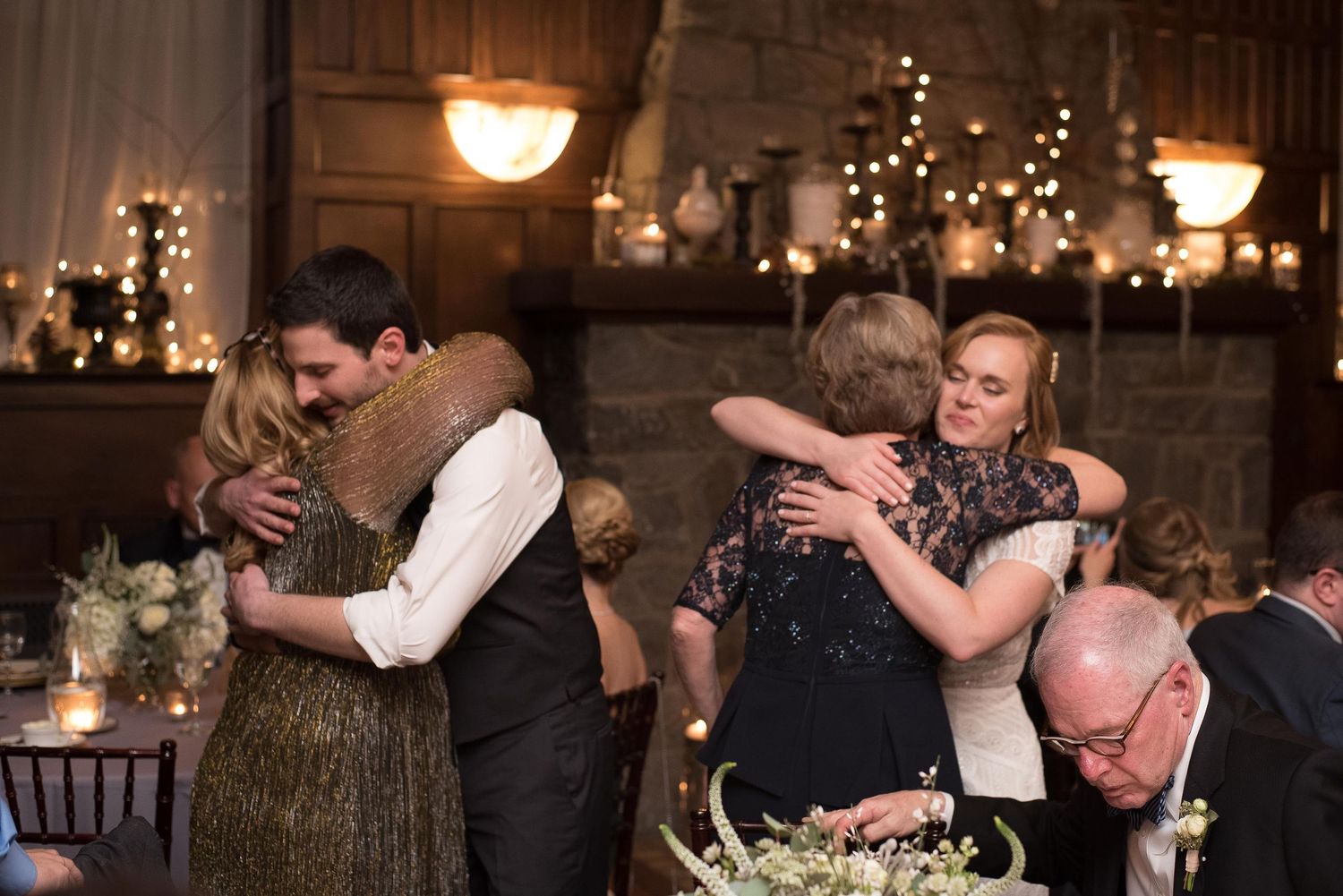 Guests sharing warm embraces at an elegant wedding reception with candlelit decor and string lights.