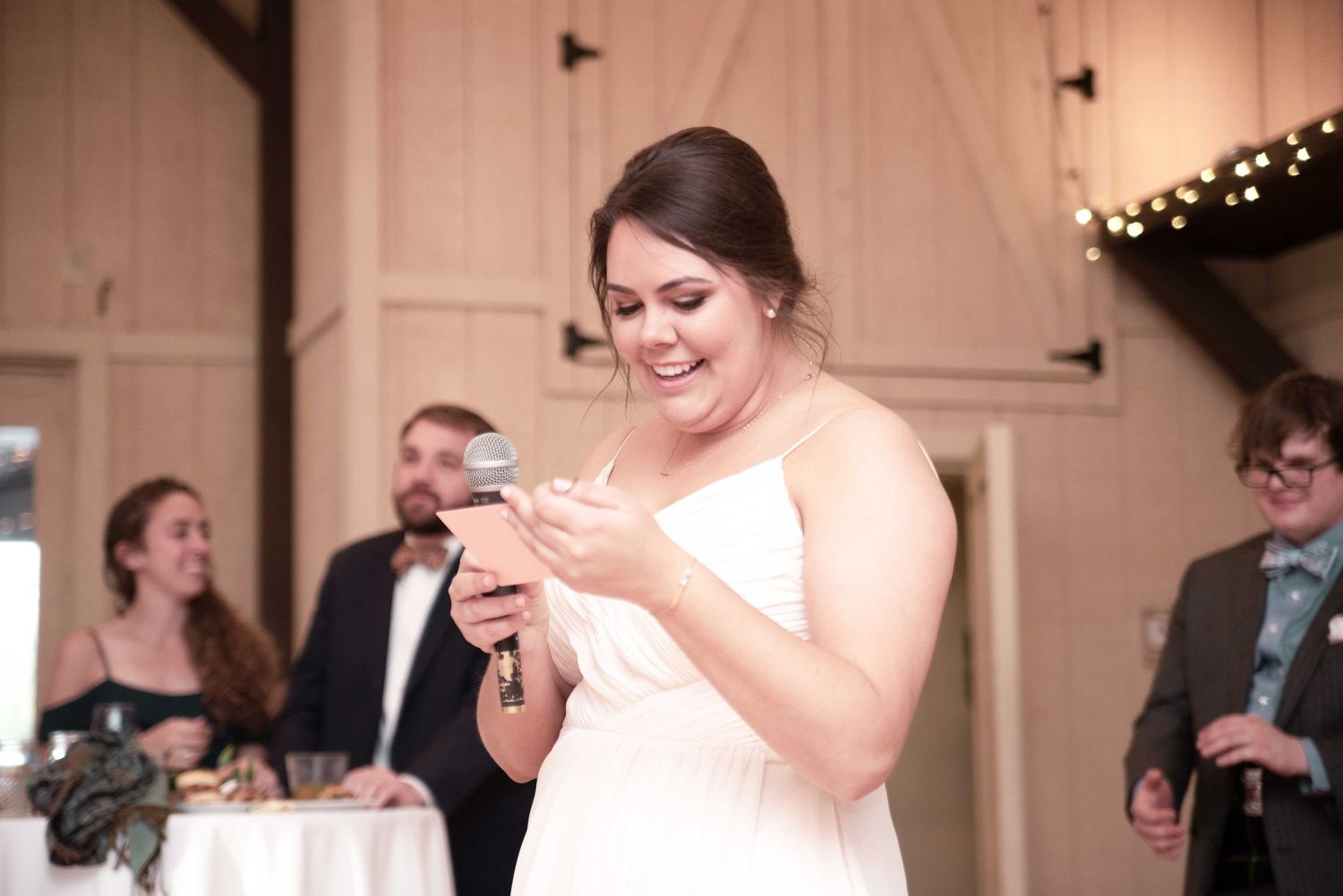 Bride in white dress smiling while giving a speech with microphone at a barn wedding reception.