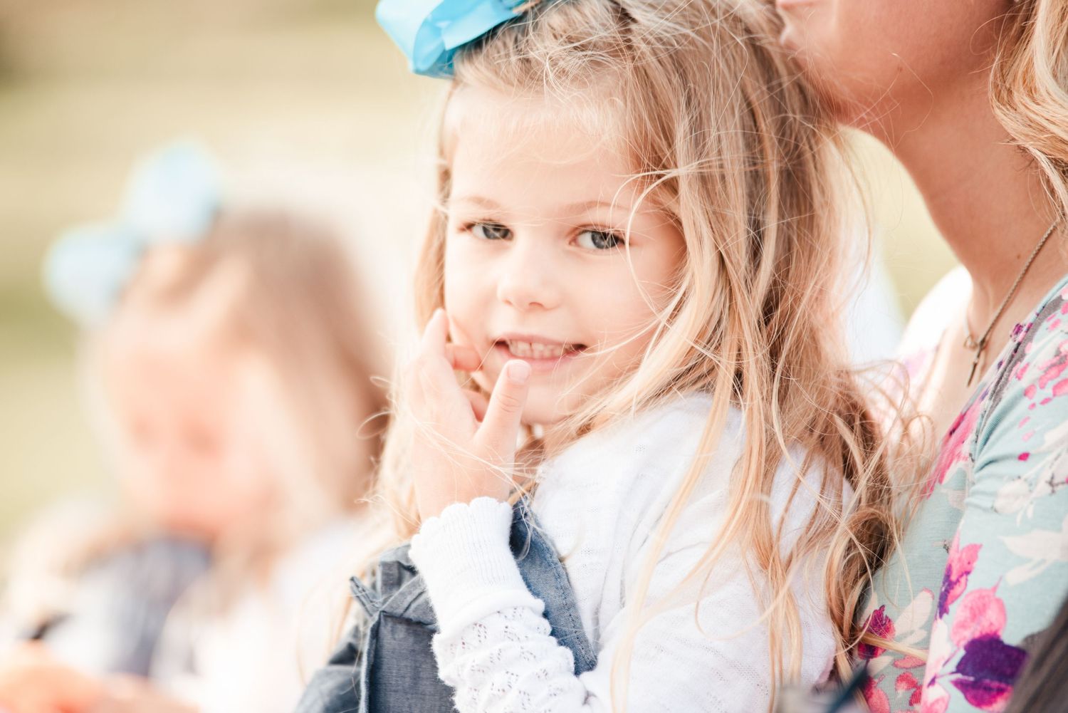 Adorable young blonde flower girl with blue bow smiles during outdoor wedding celebration.