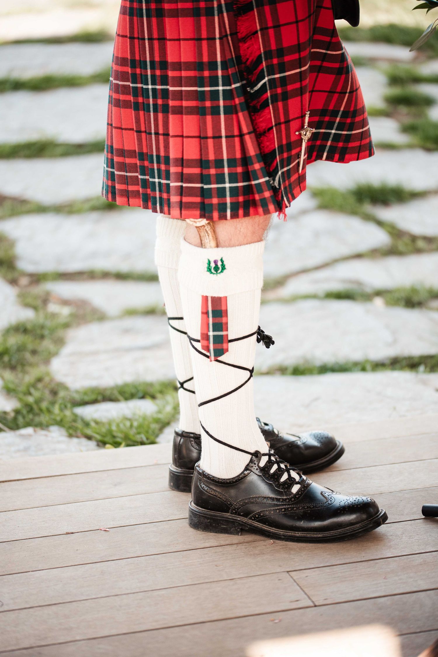 Man wearing traditional Scottish red tartan kilt with white knee-high socks and black brogue shoes on wooden deck.
