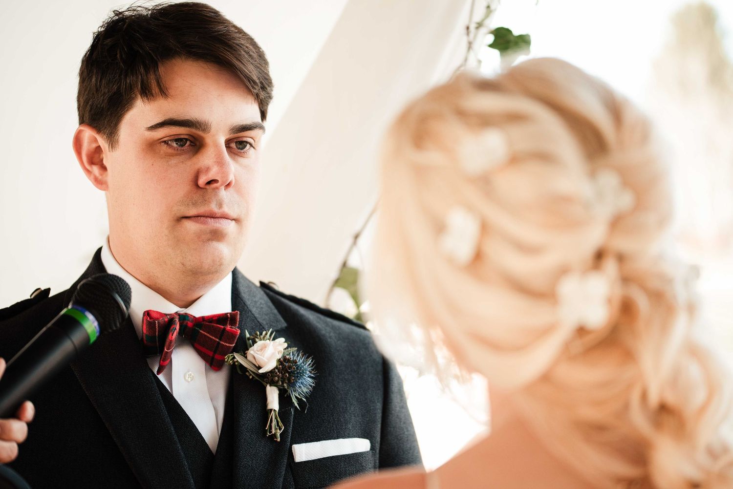 Groom in tartan bow tie and boutonniere gazes at blonde bride during wedding ceremony.