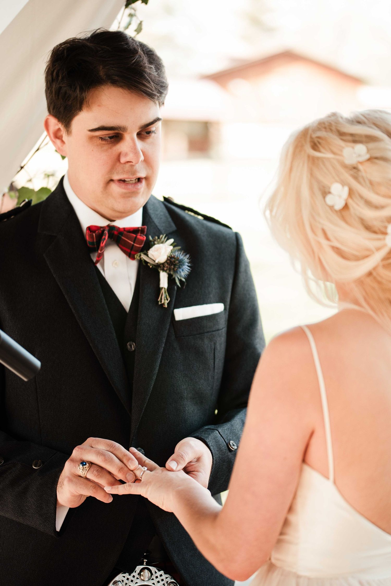 Groom in black suit and plaid bow tie placing wedding ring on bride's finger during ceremony.
