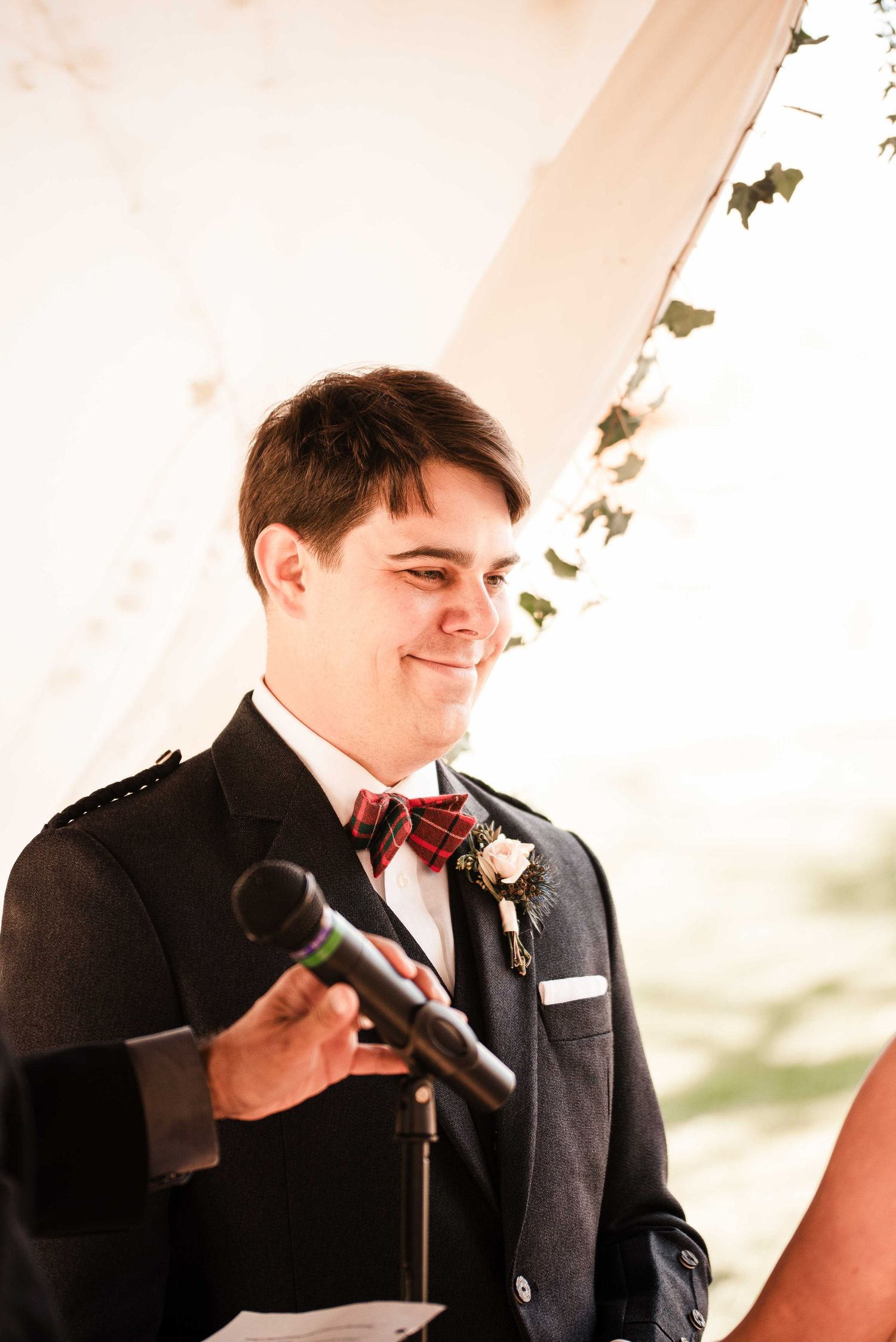 Groom in dark suit with plaid bow tie and boutonniere smiling while holding microphone at wedding ceremony.