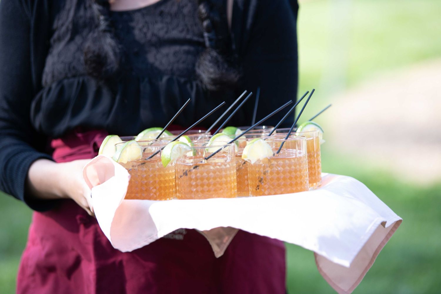 Server holding a tray of garnished cocktails with lime and black straws at an outdoor catered event.