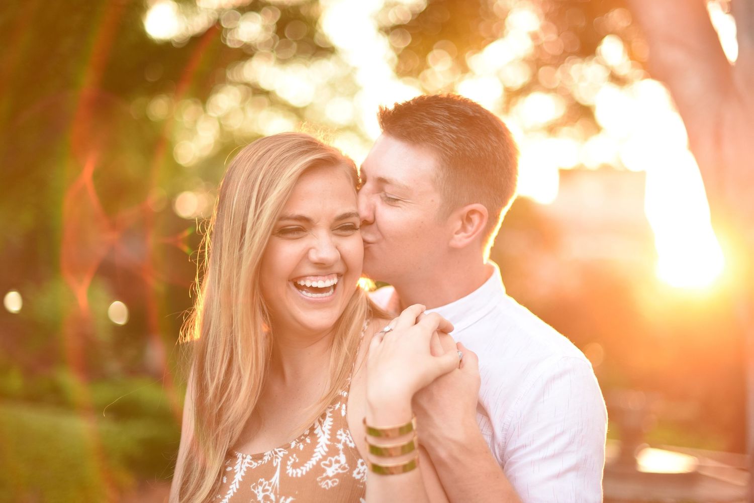 Laughing couple embracing during golden hour sunset, woman wearing gold bracelets in outdoor setting.
