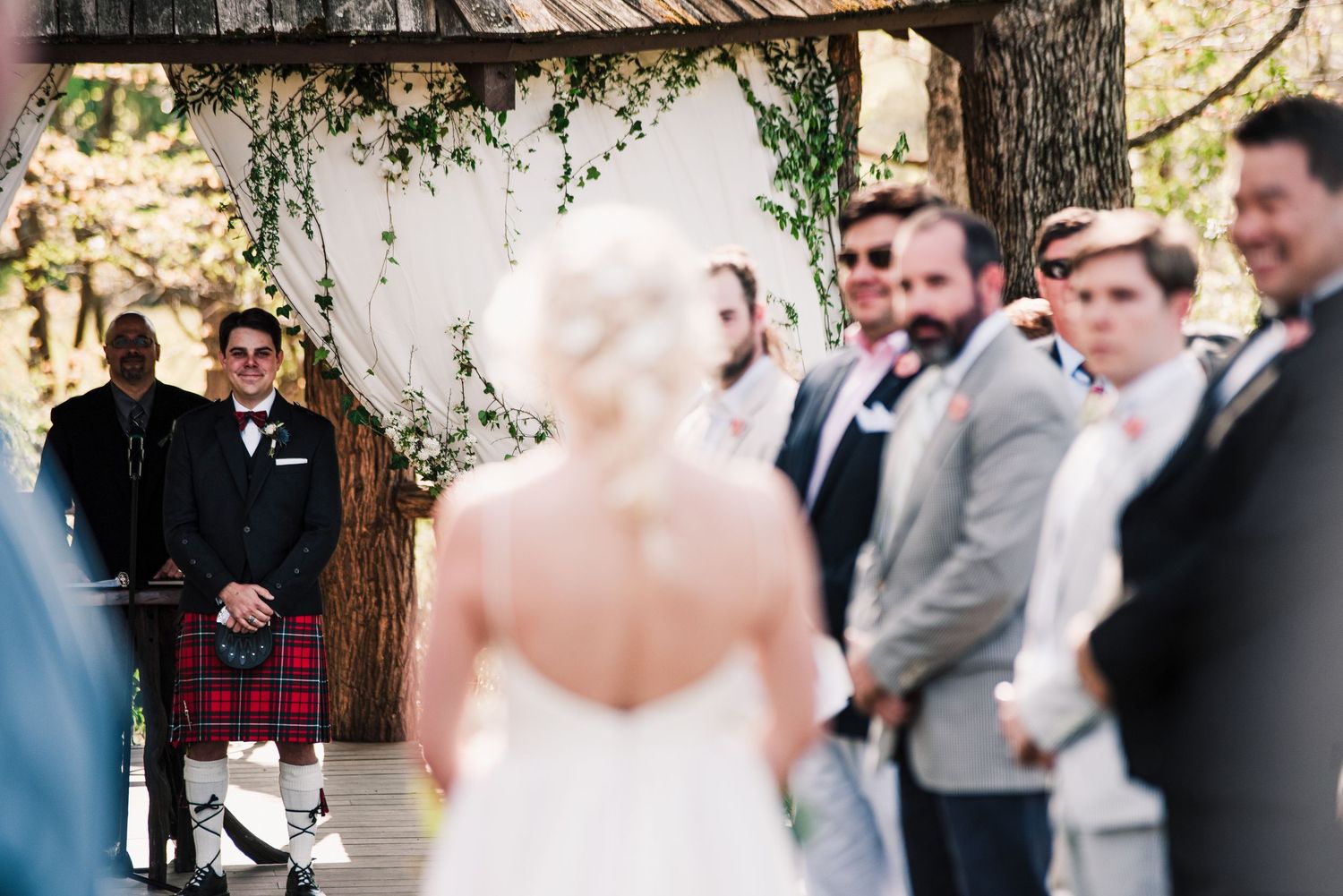 Groom in tartan kilt smiling at bride walking down the aisle at outdoor wedding ceremony.