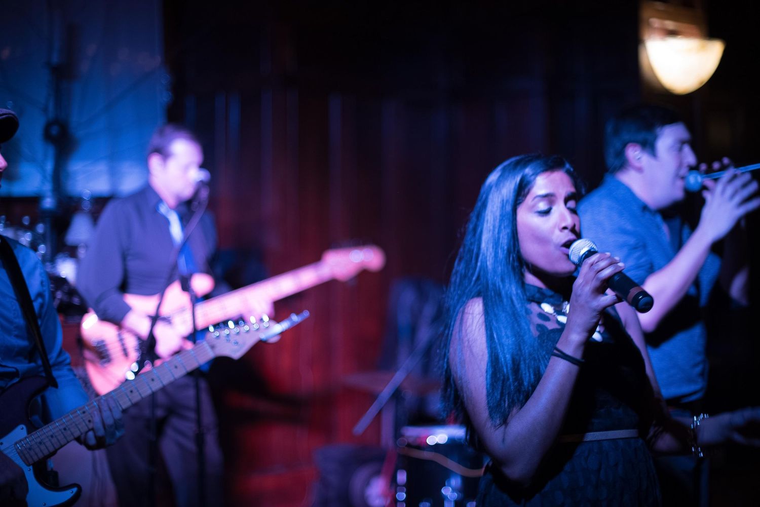 Live band performance with female vocalist singing into microphone, guitarist, and bassist on a blue-lit stage.