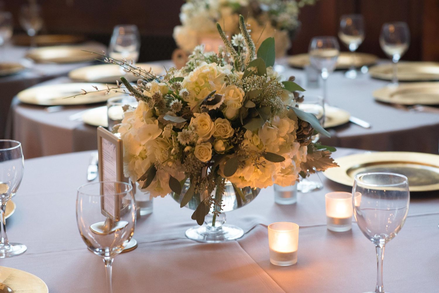 Elegant wedding centerpiece with yellow roses and greenery in a silver vase, surrounded by candles and gold-rimmed place settings.