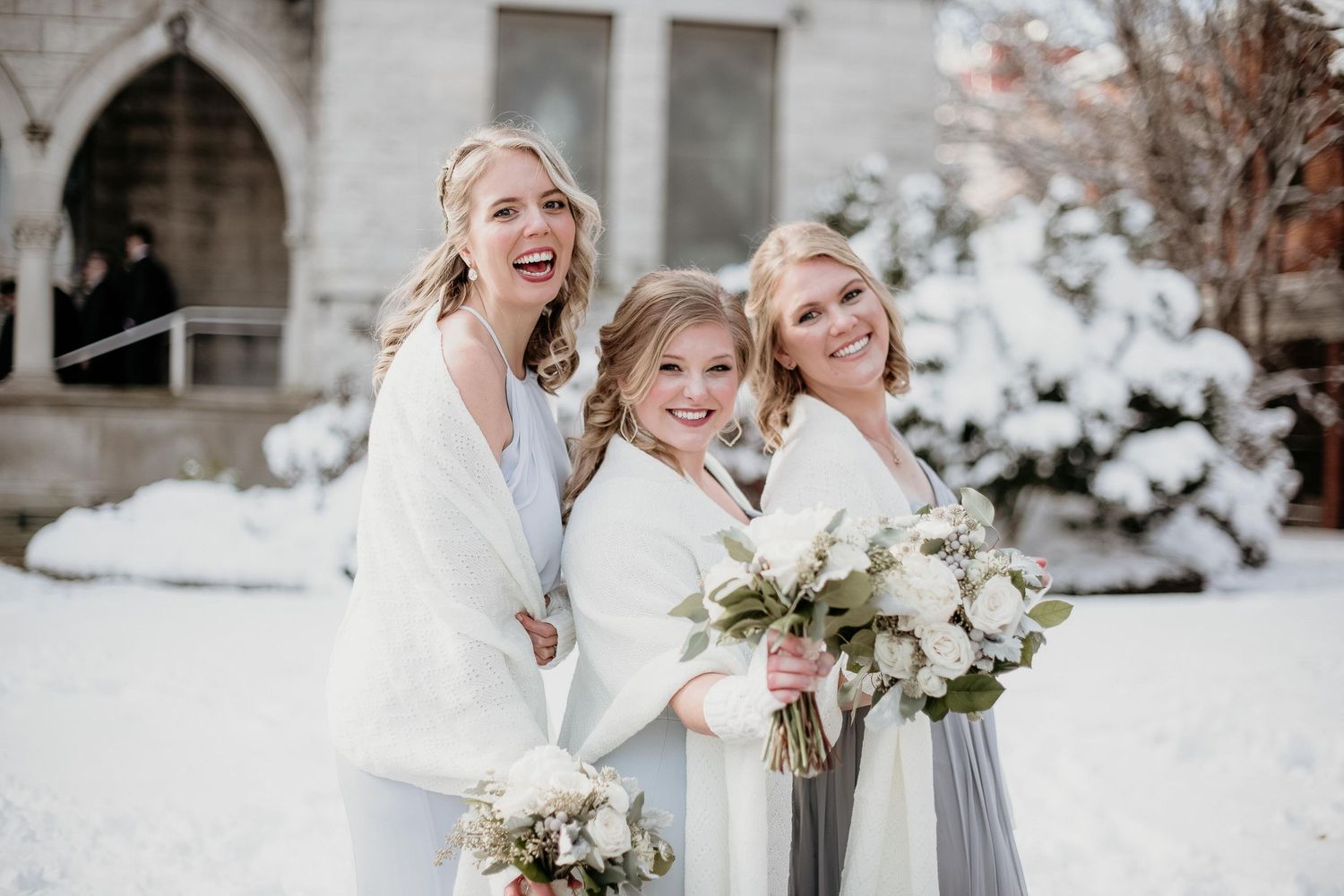 Three women in white and gray winter wedding attire smile outdoors in snow, holding white floral bouquets near a church.