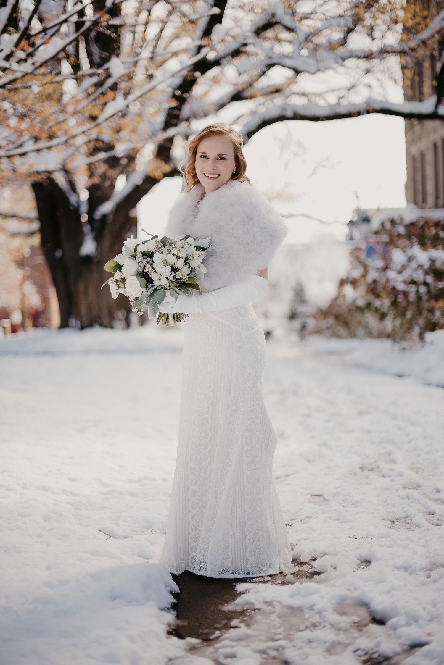 Bride in white lace gown and fur shawl holds bouquet in snowy winter outdoor setting.