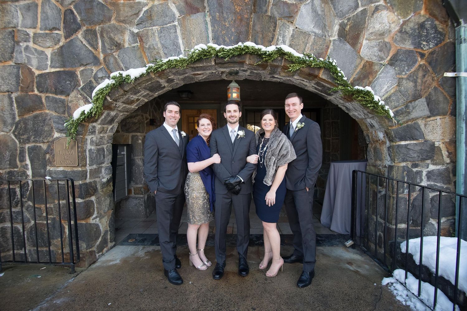 Five elegantly dressed people posing under a stone archway decorated with greenery at a formal event.