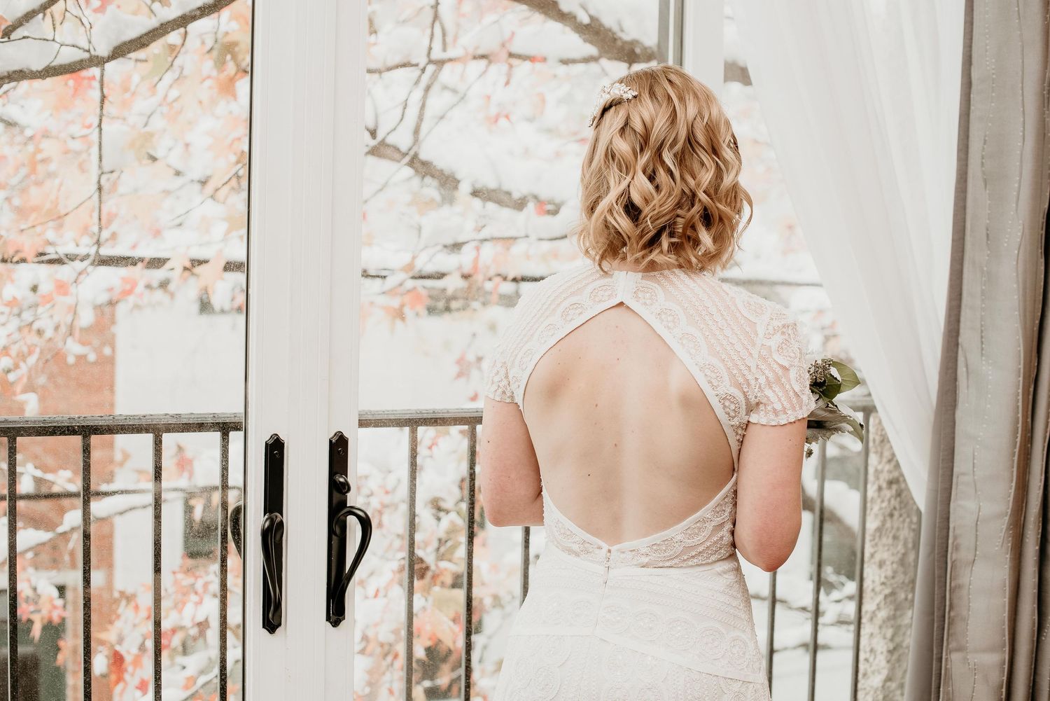 Bride in backless lace wedding dress standing at open French doors with autumn foliage in background.