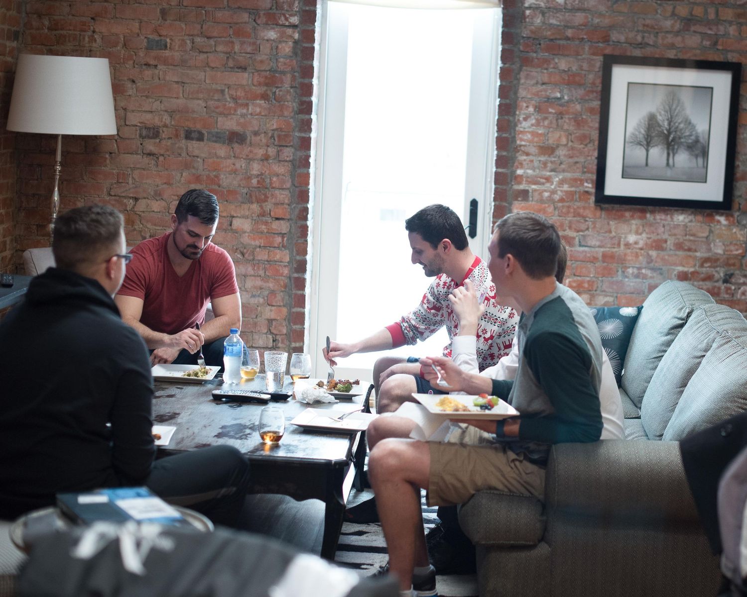 Four men sharing a meal around a coffee table in a cozy brick-walled living room with natural light.