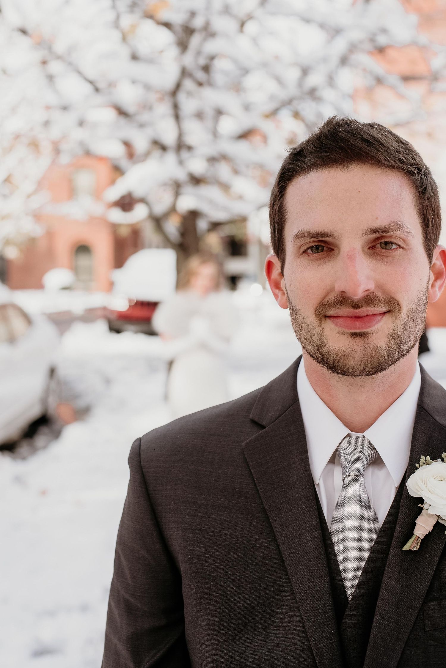 Groom in dark suit with silver tie and boutonniere smiling outdoors in snowy winter wedding setting.