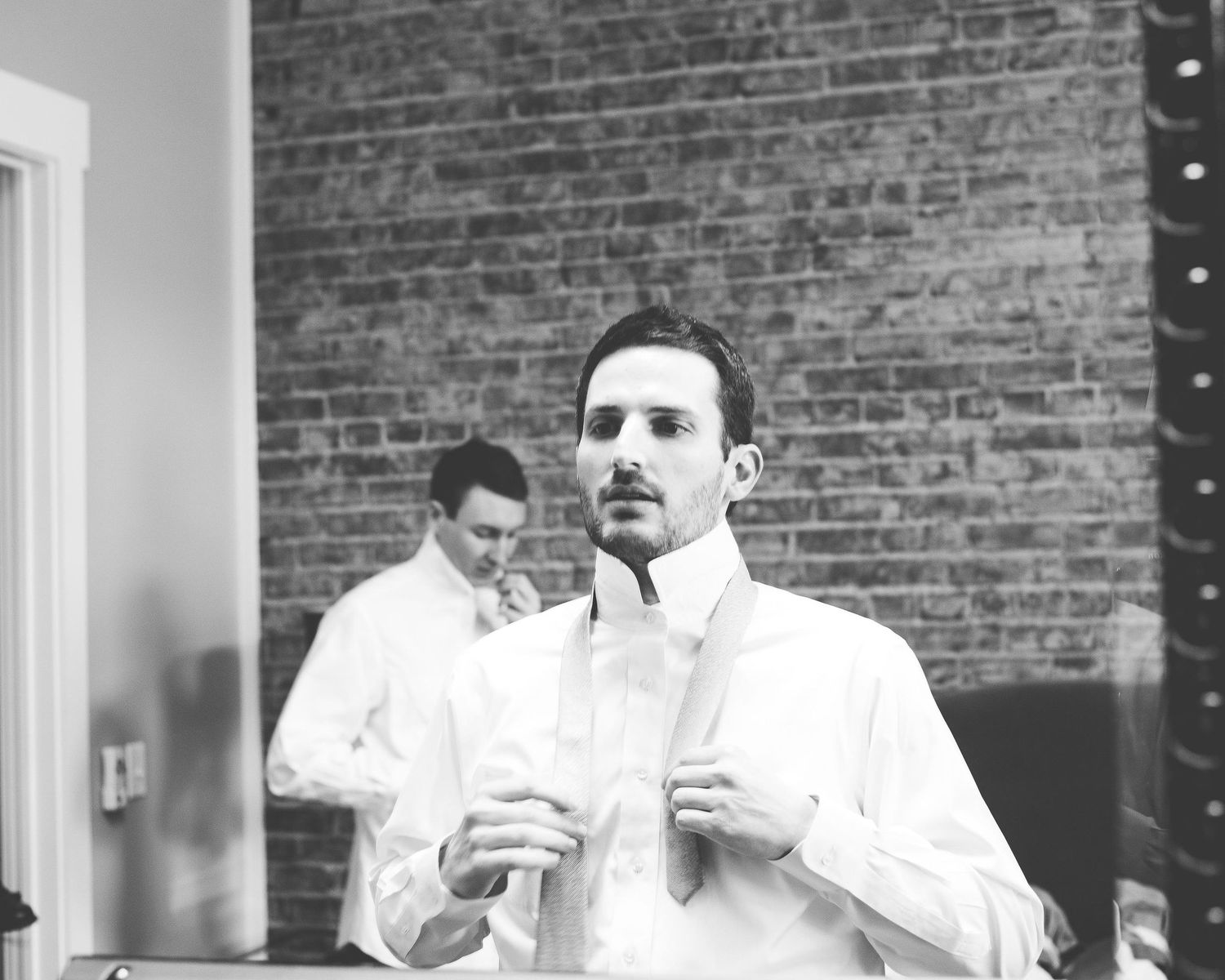 Black and white photo of a groom adjusting his shirt collar while another man stands in the background.