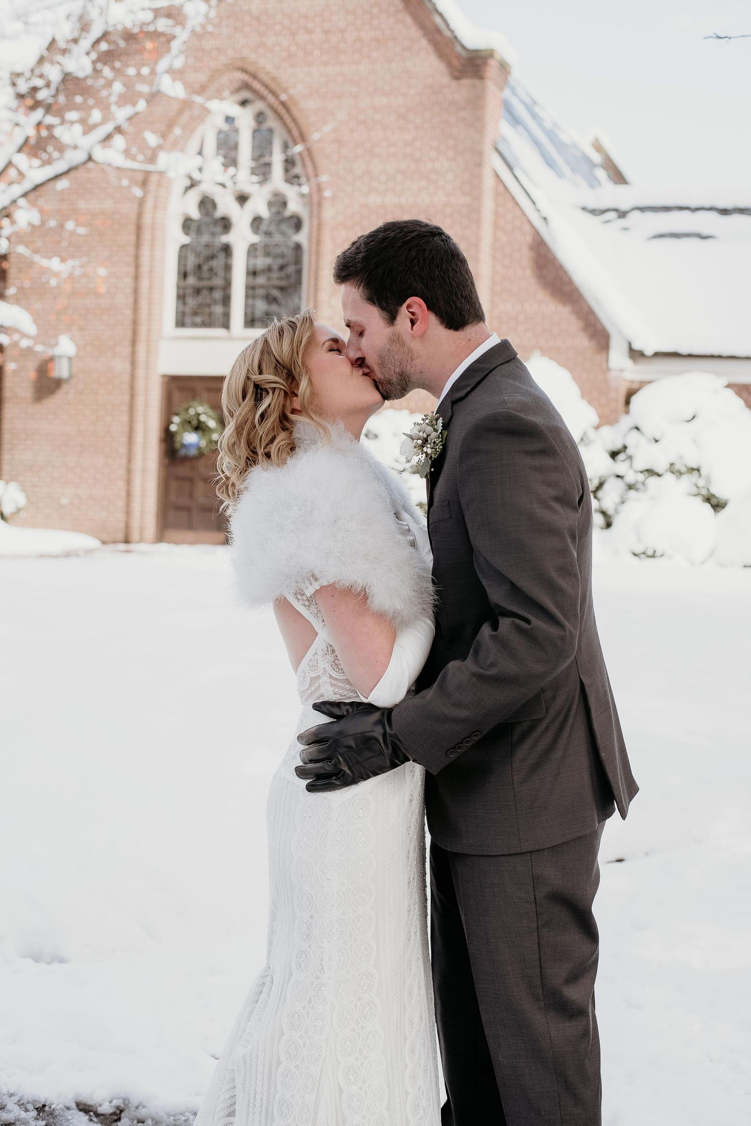 Bride and groom share first kiss outside snow-covered brick church, bride in fur wrap and groom in gray suit.