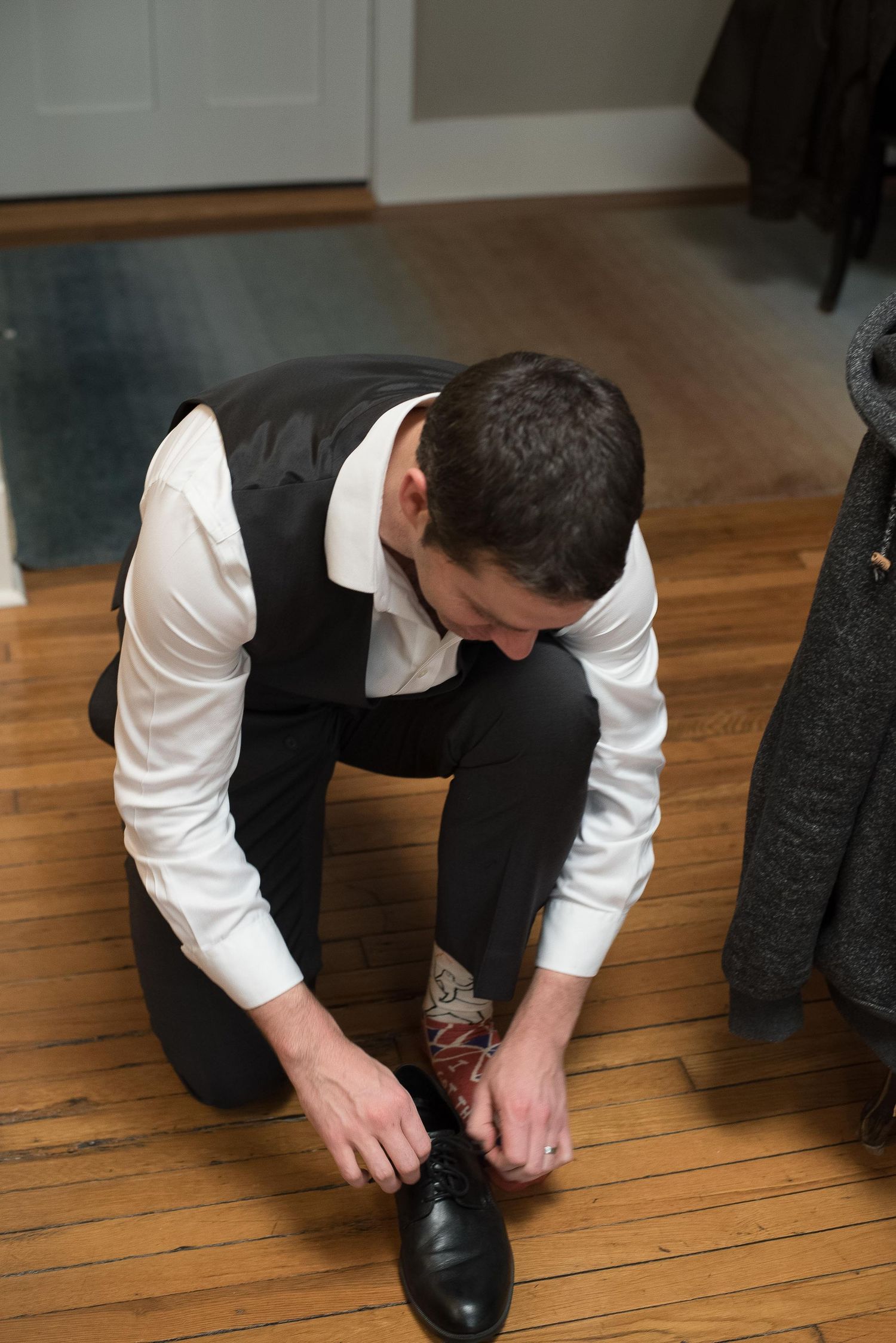 Groom kneeling on hardwood floor tying black dress shoes while wearing white shirt and black vest.