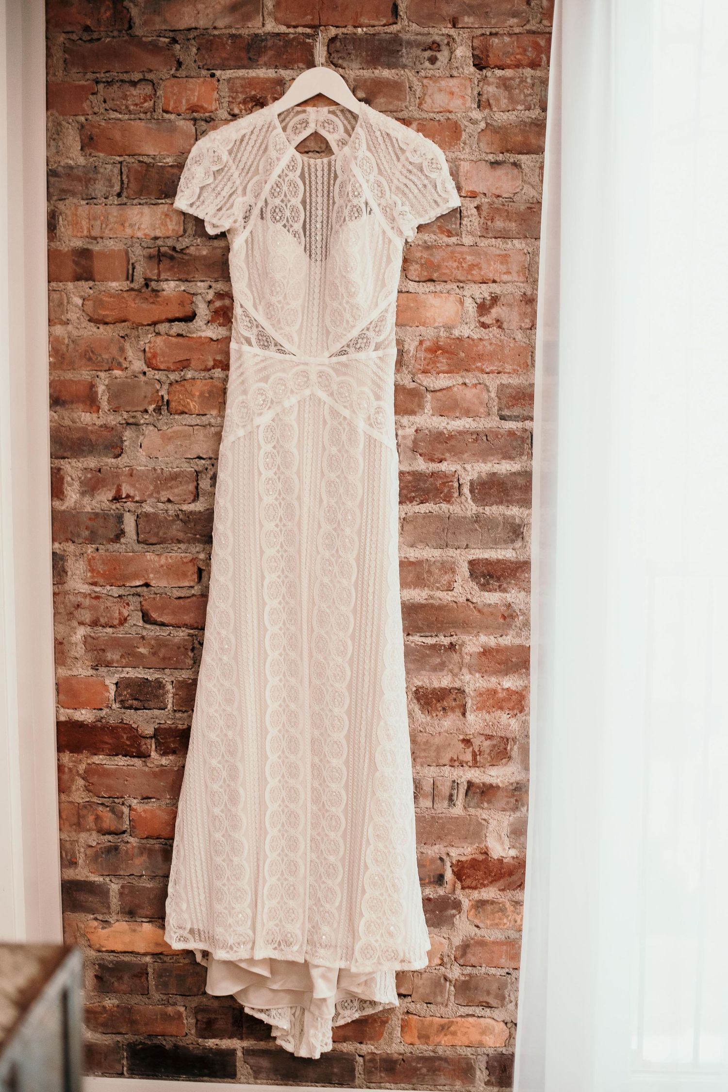 Elegant white lace wedding dress with cap sleeves hanging on a hanger against a rustic brick wall.