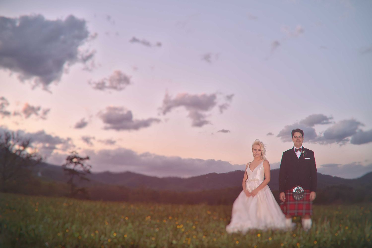 Wedding couple in pink dress and red tartan kilt pose in meadow during golden hour with mountain views.