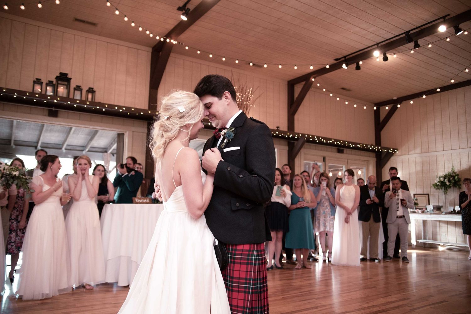 Bride and groom share first dance at rustic barn wedding reception, groom wearing traditional red tartan kilt.