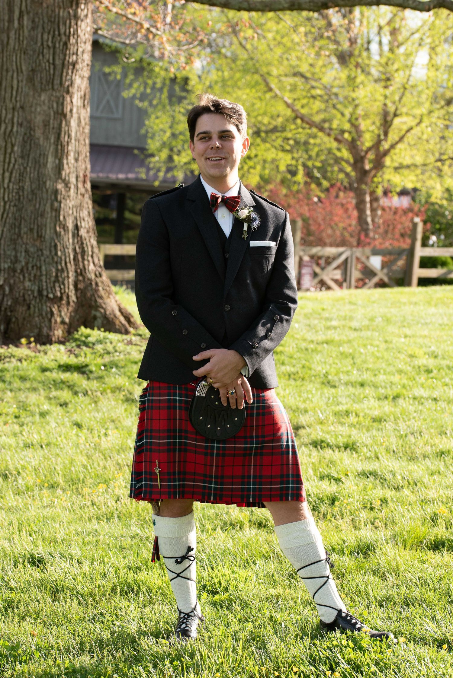 Man in traditional Scottish kilt outfit with red tartan, black jacket, bow tie, and ghillie brogues standing on green lawn.