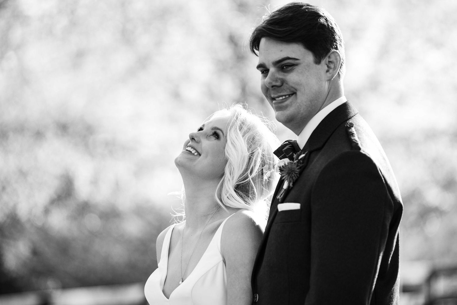 Black and white wedding photo of smiling bride and groom posing outdoors among blurred trees.