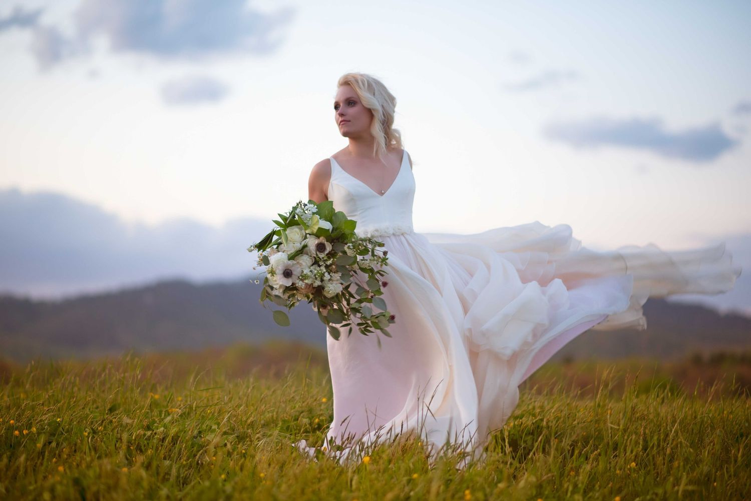 Bride in flowing white gown holding green bouquet stands in mountain meadow at sunset.