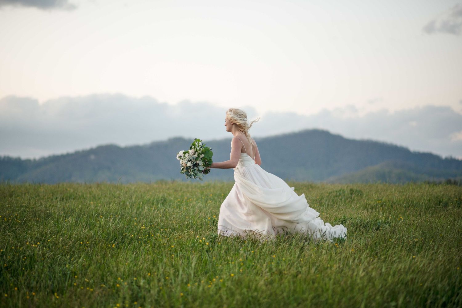 Bride in white gown running through green meadow holding bouquet with mountain backdrop.