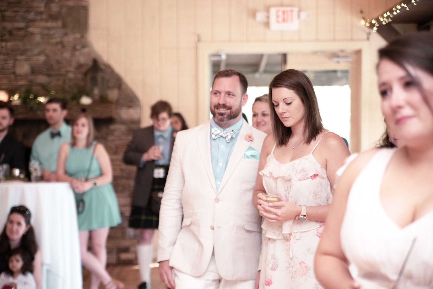 Couple in formal attire at a wedding reception, man in beige suit with bow tie, woman in floral dress holding a drink.