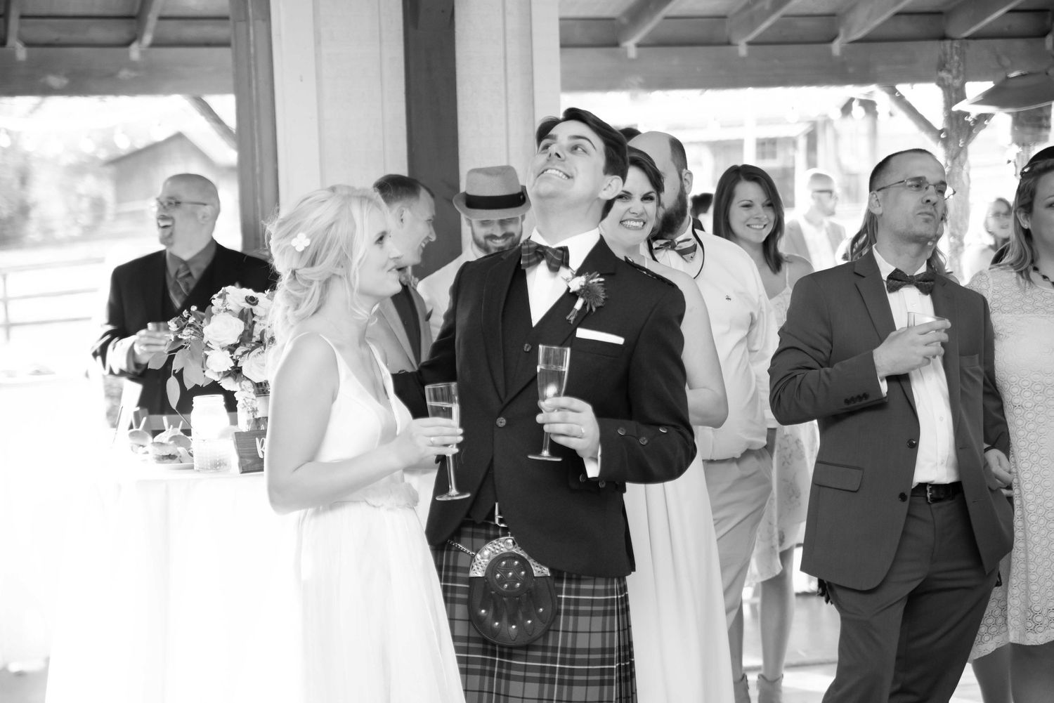 Black and white photo of bride and groom smiling during wedding reception toast with guests in background.
