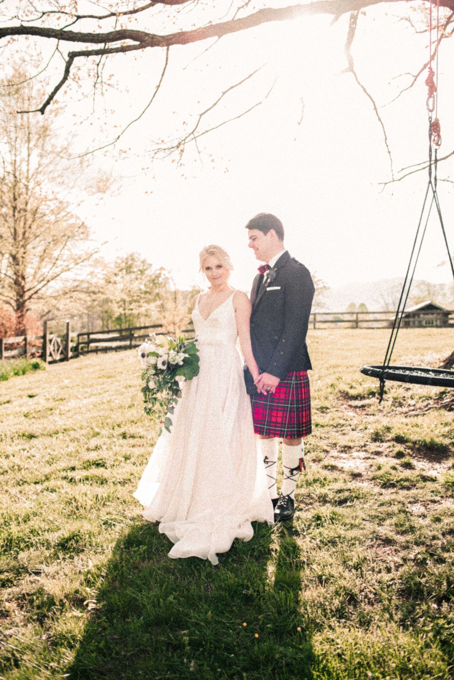 Bride in white gown and groom in red tartan kilt pose together outdoors on green grass at rustic farm wedding.